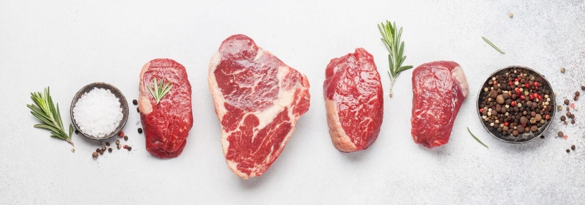 a variety of different steak cuts on white tabletop background next to sea salt and peppercorns as well as herbs